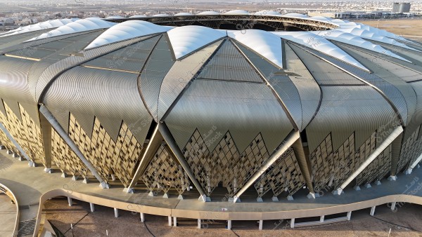 An aerial view of a stadium with a unique architectural design resembling a crescent. The roof is covered with curved white panels, surrounded by gold-colored panels on the sides. In the background, there is a view of a cityscape with multiple buildings.