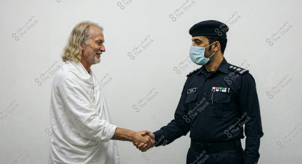 Two men are shaking hands in the image. The man on the left is wearing a loose white garment and has long gray hair. The man on the right is in a dark-colored police uniform, wearing a medical mask and a hat. His uniform features badges and military ranks. The setting appears formal due to the attire.