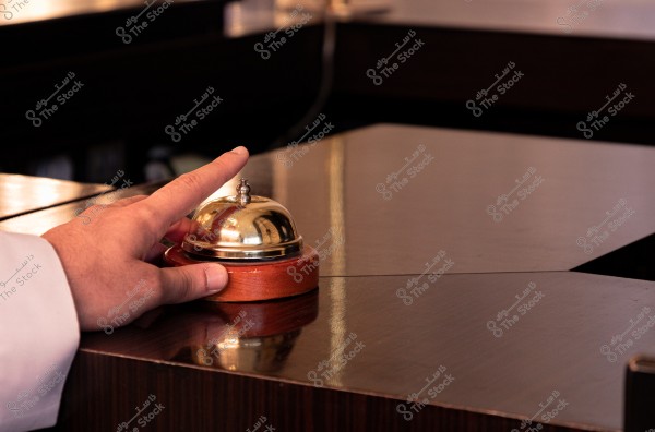 يد شخص تضغط على جرس مكتب استقبال ذهبي صغير موضوع على سطح خشبي لامع. الجرس ذو قاعدة خشبية مستديرة والجو العام للمكان يبدو كأنه مكتب استقبال في فندق أو مطعم.