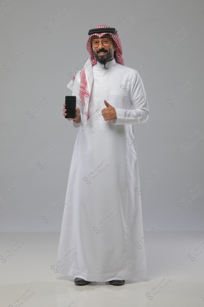 A portrait of a man wearing a white thobe and a red checkered shemagh with an agal, standing and smiling while holding a smartphone in one hand and giving a thumbs-up with the other. The background is light gray.