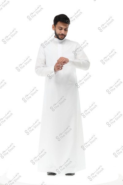 An image of a man wearing a long white thobe, resembling traditional Gulf attire, possibly from Saudi Arabia. He stands against a bright white background, looking down at his hands while adjusting the sleeves of the thobe. The outfit and colors reflect simplicity and elegance.
