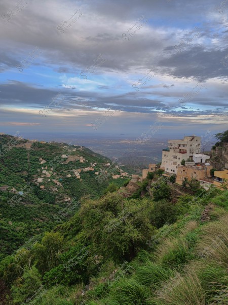 A mountainous landscape featuring green hills with houses scattered along the slopes. The horizon shows a blue sky with some clouds, and the foreground is dense with trees and greenery. Traditional buildings are constructed on the mountainside.