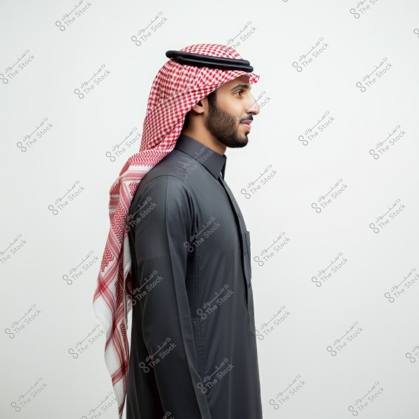 A portrait of a man from the side against a white background. The man is wearing a dark-colored thobe with a red and white checkered keffiyeh and a black agal. The style suggests the attire is from the Arabian Peninsula.