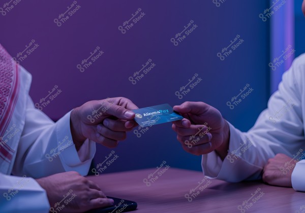 An image showing two hands, both wearing white garments, exchanging a payment card labeled \"Alinma\". One hand is wearing traditional clothing, suggesting the person might be from Saudi Arabia. The background is softly lit with purple and blue lights.