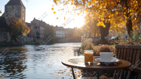 Image focusing on a wooden table placed by a river under golden autumn sunlight. On the table, there is a glass filled with orange juice and a white coffee cup with a saucer. In the background, the riverbank is lined with historic buildings and green trees with yellow and brown leaves.