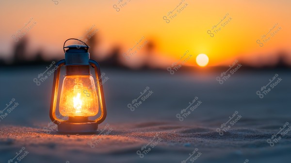 A traditional oil lamp lit on the sand during sunset. The sky features various shades of orange and yellow, with the sun setting on the horizon. The background is blurred, giving a peaceful and warm effect to the image.
