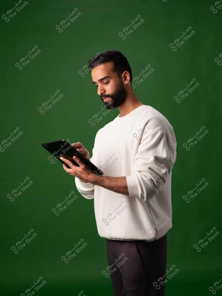 A portrait of a man wearing a white long-sleeve shirt and dark pants, standing against a green background. He appears to be focused on a tablet he is holding in his hands.