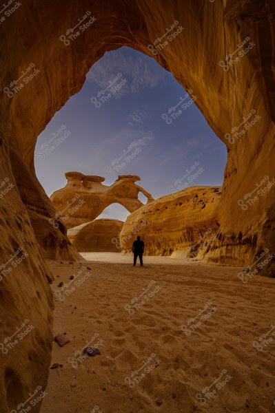 A person standing in a sandy valley surrounded by massive rock formations resembling arches under a blue sky with few clouds. The rocks are a brownish-orange color, giving a warm and contemplative feel to the natural scene.
