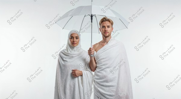 The image shows a man and a woman wearing traditional white Ihram clothing. The man on the right is holding an open white umbrella, and the woman is standing beside him, wearing a white hijab. The background is simple and white, highlighting the details of the clothing. The image suggests preparation for performing Hajj or Umrah.