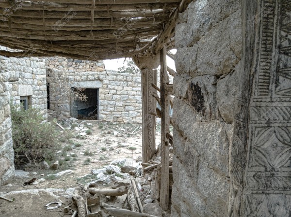 An image of an old stone building showing a section of a decorated wall and a roof made of wooden sticks. The ground appears neglected, covered with rocks and sparse grass. There is an opening for a door in the background wall with dry plants around it. The overall feel suggests an early abandoned setting or a historical village.