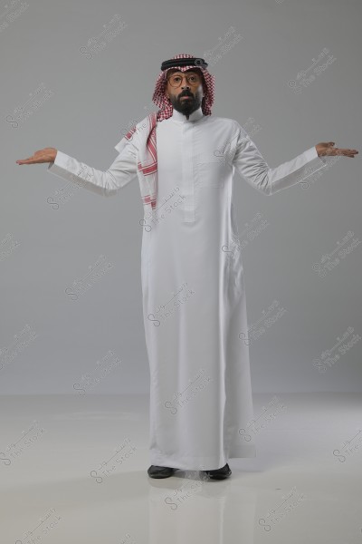 A portrait of a man in traditional Saudi attire, wearing a white thobe, white and red checkered ghutra, and black agal. He stands with his arms raised to the sides, suggesting a gesture of inquiry or questioning, set against a simple gray background.