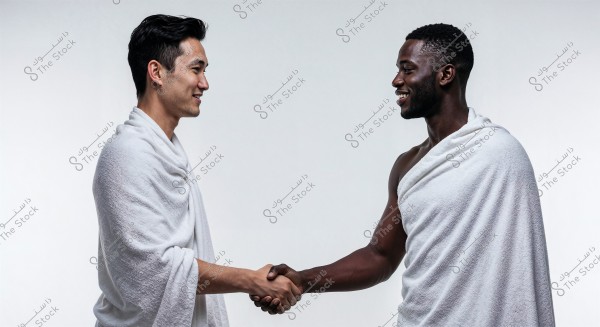 The image shows two men, one of Asian descent and the other of African descent, shaking hands with a smile. Both are wearing white garments similar to Islamic ihram attire, which consists of simple white cloth wrapped around the body. The background is plain white.