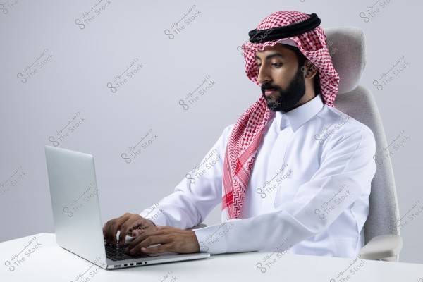An image of a man wearing a thobe, ghutra, and agal, sitting on a chair and working on a laptop. The background is light gray.