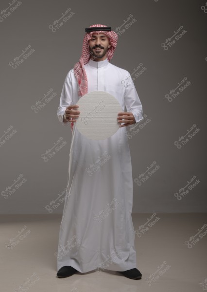 A portrait of a man wearing traditional Saudi attire, consisting of a white thobe with a red headscarf (shemagh) and a black agal. The man is smiling and holding a flat, circular white object. The background is simple and neutral-colored.