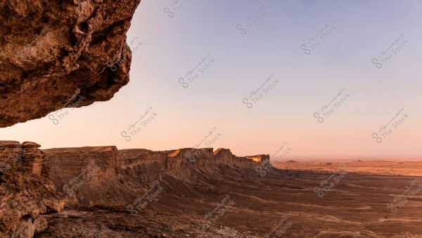 A natural landscape featuring a stretch of rocky cliffs in a desert area under a light blue sky during sunrise or sunset. The prominent mountain rocks extend dramatically across the scene, showing gradients of brown and yellow blending with the soft sunlight.