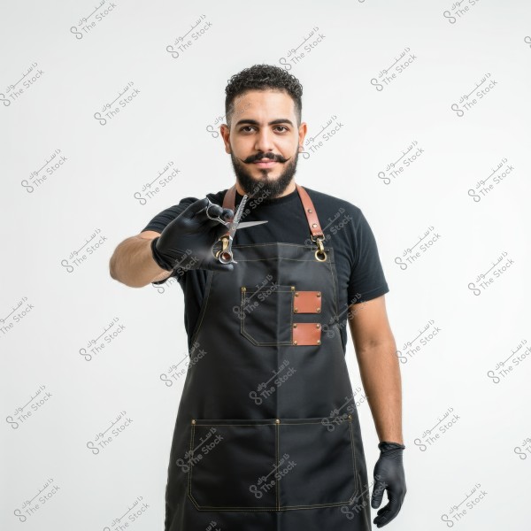 A portrait of a bearded man wearing a black apron and gloves, holding a hair cutting scissors toward the camera. He appears to be a professional barber, and the apron features brown leather details. The background is entirely white.