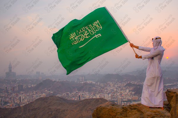 A snapshot of a Saudi man wearing the traditional Saudi dress, carrying the Saudi flag over a mountain in the Makkah Al-Mukarramah region, and the sky appears clear, the royal clock tower building in the Grand Mosque, buildings and landmarks, the Grand Mosque of Mecca.