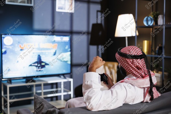 A man wearing traditional thobe and keffiyeh is sitting on a sofa, holding a video game controller. The screen in front of him displays a game with the text \"You Win!\", indicating he has won the game. The room is lit by a floor lamp and contains shelves with some decorative items.
