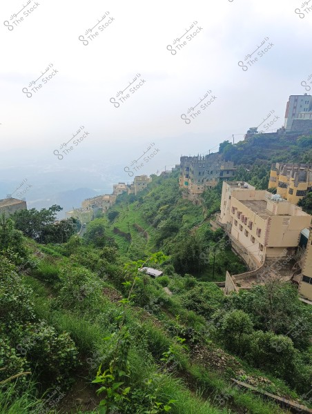 The image shows a mountainous landscape with greenery and houses built on the slopes. The buildings are scattered along the hillside and have a simple design with various colors. The horizon appears misty, suggesting humid or rainy weather. The view from above features various shades of green from trees and plants.