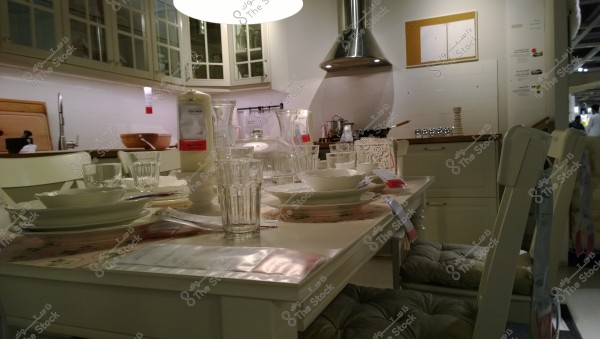 Image of a modern kitchen with simple design elements. The table is set with dishes and glass tumblers, alongside a large square candle and serving bowls. White upper cabinets with glass doors reveal some of their contents. A stainless steel vent hood is mounted above the stove. The kitchen appears clean and organized.