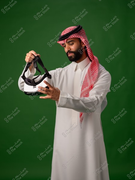 رجل يرتدي الزي السعودي التقليدي يقف على خلفية خضراء وهو يحمل نظارة واقع افتراضي بيضاء بيديه وينظر إليها بتركيز.

-
