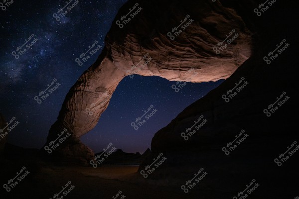 Image of a massive rock arch in the desert under a starry night sky. The arch is softly lit along its edges, giving the image depth and a natural beauty.