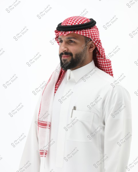 A portrait of a man wearing a white thobe and a red and white checkered ghutra with a black agal. He stands with a calm expression on his face. The background is white, making the image stand out more clearly. The attire is traditional in Saudi Arabia.