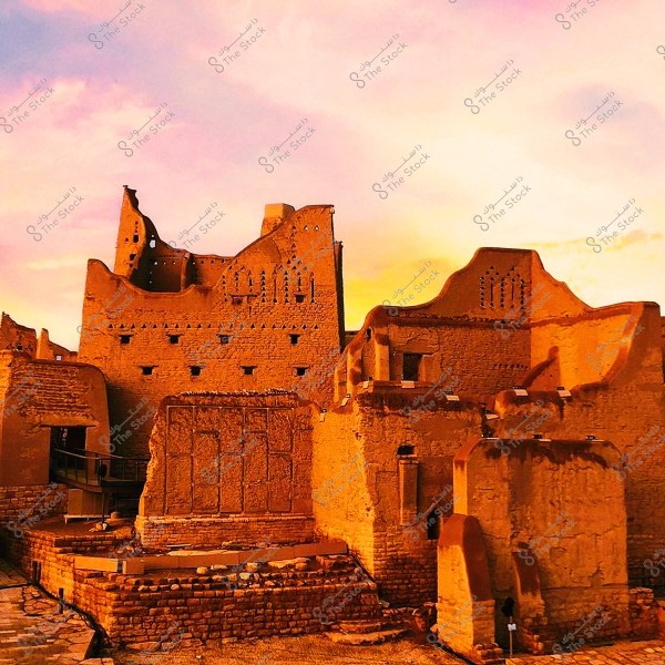 Image showing a view of ancient mud-brick buildings in the historic area of Diriyah, Saudi Arabia, under a sky with warm sunset colors. The traditional architecture features mud walls adorned with Arabic designs and patterns.