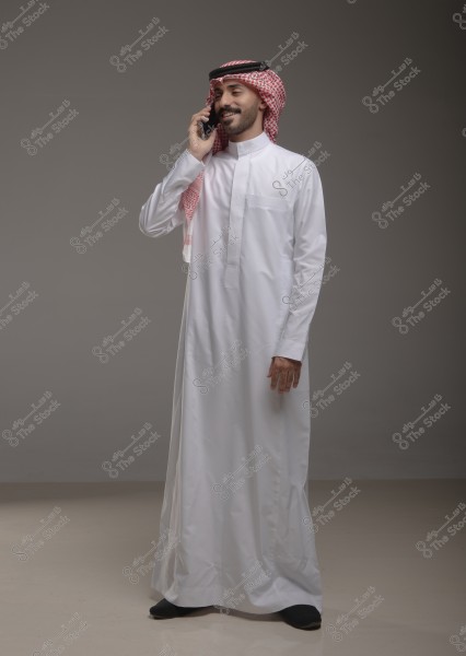 A man wearing traditional Saudi attire, consisting of a white thobe, a red checkered shemagh, and a black agal, stands against a grey background talking on the phone with a smile.