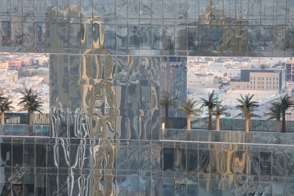 Image of a glass building reflecting sunlight with distorted reflections of palm trees, set against a backdrop of a cityscape featuring various buildings and structures. The palm trees add an aesthetic touch to the urban scene.
