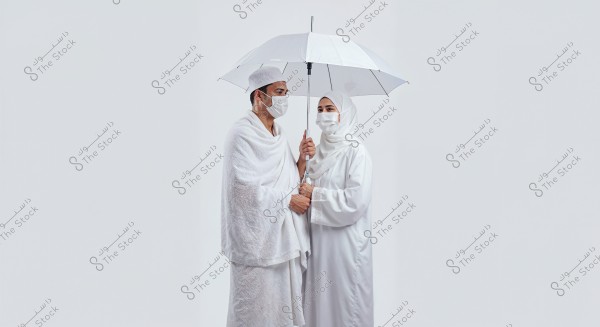 A portrait of a couple wearing traditional Ihram clothing for Hajj, standing under a white umbrella. The man is dressed in a white wraparound and shawl, while the woman wears a white abaya and headscarf. Both are wearing medical face masks. The background is plain white.