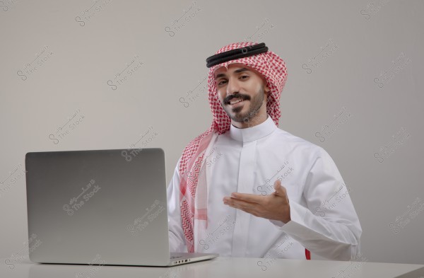 صورة لرجل يرتدي الزي السعودي التقليدي, مكون من الثوب الأبيض والغترة الحمراء مع العقال الأسود, جالس أمام جهاز كمبيوتر محمول. تبدو عليه علامات التواصل أو الحديث بإشارة من يده. خلفية الصورة محايدة.