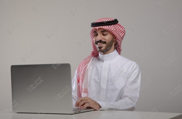 A portrait of a man sitting in front of a laptop wearing traditional Saudi attire, including a white thobe, red checkered headscarf (ghutra), and black agal. He is smiling and focused on the screen. The background is neutral.