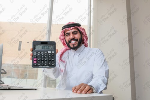 رجل سعودي يرتدي الزي السعودي التقليدي يعمل في مكتب بواجهة زجاجية ويستخدم آلة الحاسبة أثناء العمل