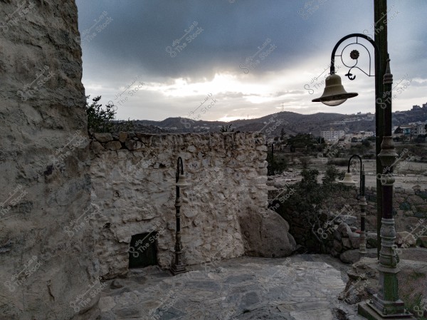 An image depicting a natural scene with an old stone wall next to a traditional-style lamp post. The background reveals a range of hills with a few residential buildings scattered along the slopes. The sky is overcast with clouds and a subtle appearance of either sunrise or sunset.