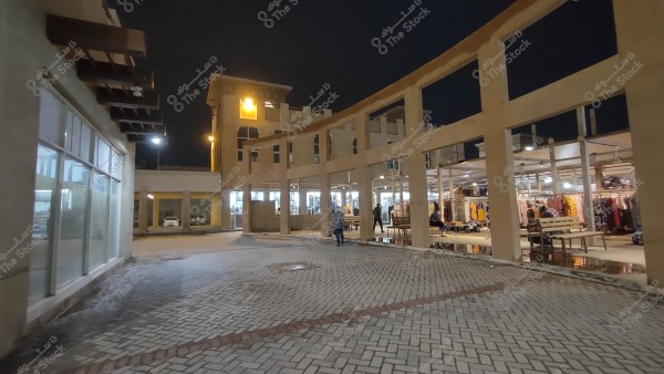 The image shows an outdoor market scene at night, with a multi-story building lit brightly in the background. Shops line a walkway displaying various clothing and merchandise. The ground is covered with interlocking tiles, and several people are walking or sitting on benches around the market.