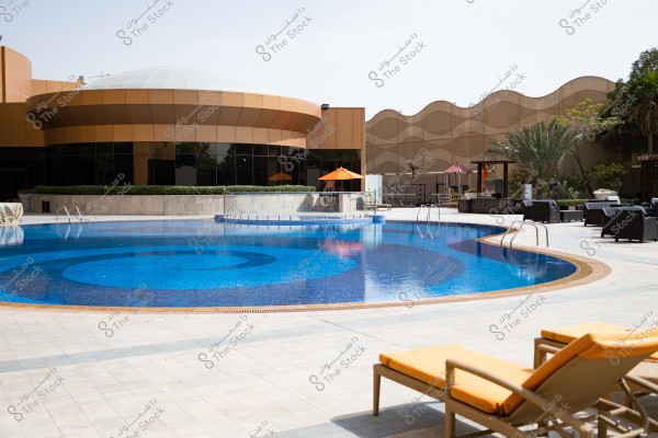 The image shows a circular swimming pool surrounded by a modern building with a wavy design in orange and cream colors, with comfortable lounge chairs near the pool edge and orange umbrellas. In the background, there are plants and palm trees enhancing the aesthetic of the area.
