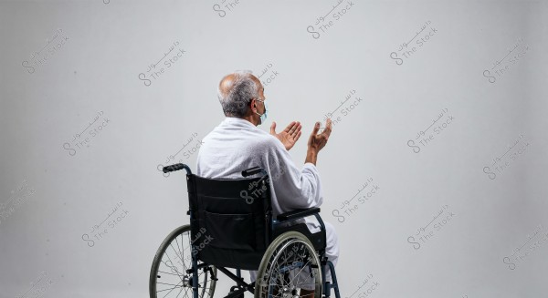 The image shows an elderly man sitting in a wheelchair, wearing a white robe and a medical mask, looking to the right while raising his hands in a prayer gesture. The background is plain white, and the man\'s features are not fully visible.