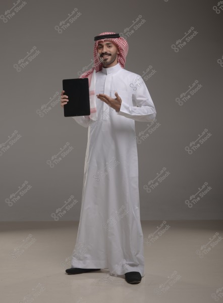 An image of a man wearing traditional Saudi attire with a white thobe, a red-checkered shemagh, and an agal. The man is smiling, gesturing with one hand, and holding a tablet with the other. The background is neutral in color.