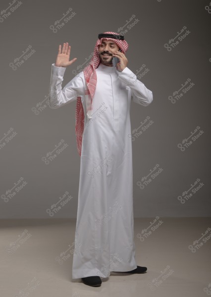 A portrait of a Saudi man wearing a white thobe and a red shemagh with a black agal. He is standing, smiling, and holding a phone to his ear with his right hand while raising his left hand as if waving. The background is plain and featureless.