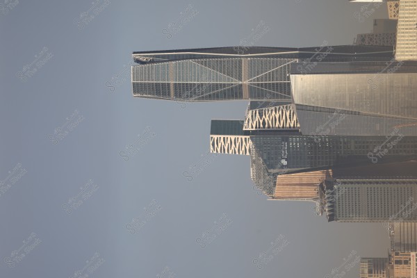 The image shows a cluster of skyscrapers in Riyadh, Saudi Arabia. Some buildings feature modern and unique architectural designs with glass facades reflecting light. The background is a clear blue sky.