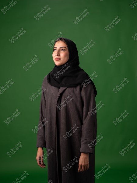 A portrait of a young woman wearing a black hijab and a dark-colored abaya, standing against a green background. She appears to be smiling and looking to her right.
