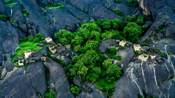 صورة جوية لقرية غيه تُظهر منازل حجرية قديمة معلقه على سفح جبل بين تكوينات صخرية ضخمة مغطاة بالخضرة. تنتشر الأشجار الكثيفة والنباتات الخضراء حول المنازل وعلى الصخور, مما يضفي مظهراً طبيعياً ومميزاً على المنطقة.