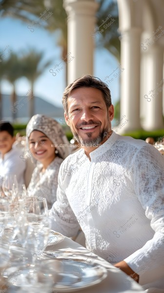 Image of a smiling man sitting at a table covered with a white cloth and glassware. The man is wearing a white garment with geometric designs. In the background, two other people are seated outside with palm trees and white columns visible.