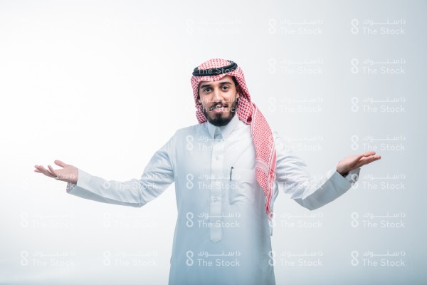 بورتريه رجل سعودي يقف على خلفية بيضاء يرتدي الثوب ويبتسم. صورة شخصية لرجل سعودي يقف بتعجب