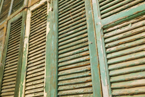 A close-up image of old wooden shutters in a faded green color. The shutters are peeling and show signs of aging and wear. Natural light highlights the details of the weathered wood and chipped paint.
