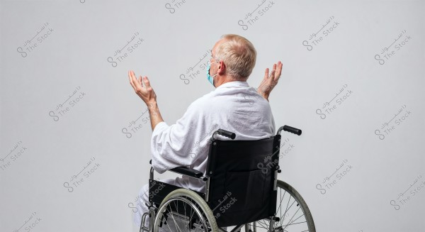 Image of a man sitting in a wheelchair, wearing a white robe and raising his hands as if in prayer. The man is seen from behind and is wearing a medical face mask. The background is grey and neutral.
