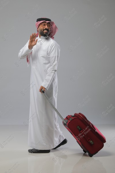 A man is wearing traditional Saudi attire with a red-checkered headscarf and a white robe. He is smiling and waving while pulling a red suitcase with a handle. He is also wearing glasses and black shoes. The background is gray with a reflective floor.