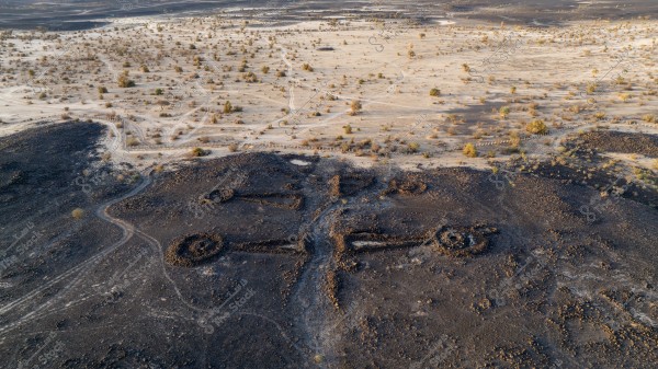 Aerial view of ancient stone structures shaped like a key in a desert area. The surrounding terrain varies between dark brown and black with sparse patches of vegetation scattered in the top part of the image. The scene indicates the presence of an ancient civilization and artistic use of the land.