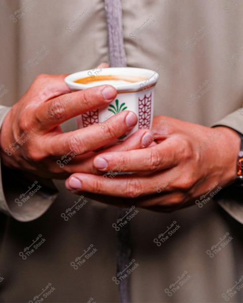 The image shows hands holding a traditional Arabic coffee cup decorated with green and brown patterns. The person is wearing a beige traditional garment, with a watch on the wrist, suggesting a traditional Arabic setting.